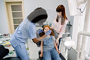 Young female African American dentist with dental tools, making dental check up of her little patient girl. Young mother