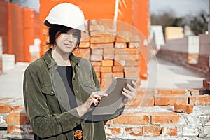 Young female architect with tablet checking blueprints against of building new modern house. Stylish happy woman engineer in hard