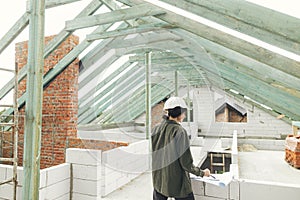 Young female architect with tablet checking blueprints against wooden roof framing of modern farmhouse. Stylish woman engineer in