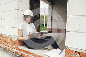 Young female architect with tablet checking blueprints while sitting in window of new modern house. Stylish woman engineer in hard