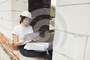 Young female architect with tablet checking blueprints while sitting in window of new modern house. Stylish woman engineer in hard