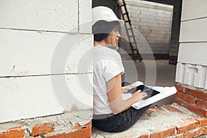 Young female architect with tablet checking blueprints while sitting in window of new modern house. Stylish woman engineer in hard