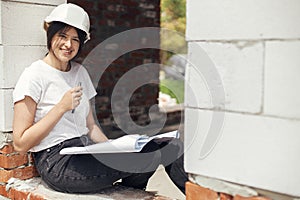 Young female architect with tablet checking blueprints while sitting in window of new modern house. Stylish woman engineer in hard