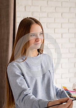 Young female artist painting on the canvas in the studio holding a brush and a palette with watercolors