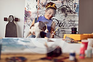 A young female artist is playing with her dog while working on a new painting in the studio. Art, painting, friendship, studio