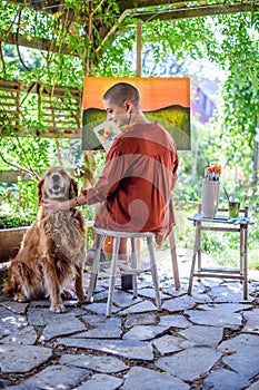 Young female artist working on her art canvas painting outdoors in her garden with golden retriever keeping her company.