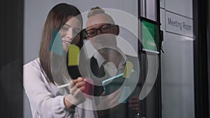 Young female assistant writing with pen on adhesive notes on glass door as male CEO talking standing in meeting room