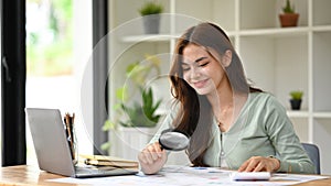 Young female auditor inspecting financial document with a magnifying glass at office desk