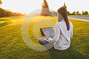 Young female back view. Woman working on laptop pc computer with blank black empty screen to copy space in city park on