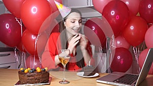 Young female blogger in birthday hat talking to laptop holding piece of cake with candle. Happy birthday woman alone at home in on