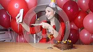 Young female blogger in birthday hat talking to mobile phone holding piece of cake with candle. Happy birthday woman alone at home