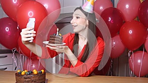 Young female blogger in birthday hat talking to mobile phone holding piece of cake with candle. Happy birthday woman alone at home