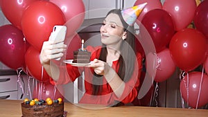 Young female blogger in birthday hat talking to mobile phone holding piece of cake with candle. Happy birthday woman alone at home