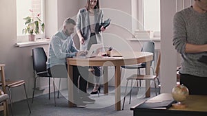 Young female boss in formal clothes comes up, sits down by male employee working with laptop to help in modern office.