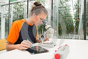 Young female botanist examining samples of plant under microscope while using digital tablet at table photo