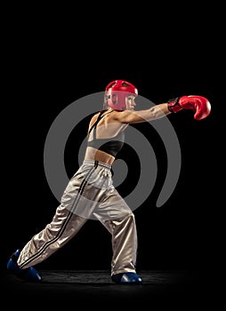 Young female boxer in boxing gloves and helmet training isolated on dark studio background. Sport, achievements