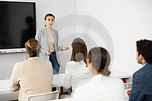 Young female business coach explaining topic while standing against group of people sitting at desks in front of her