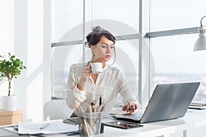 Young female business person working in office using laptop, reading and searching information attentively, drinking