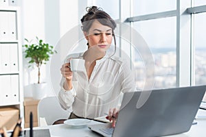 Young female business person working in office using laptop, reading and searching information attentively, drinking