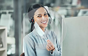 Young female call center agent smiling and wearing a headset and mask working on a computer in an office at work