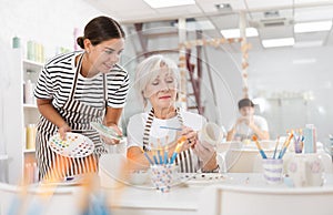 Young female ceramicist teaching elderly woman to paint ceramics in pottery studio