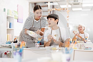 Young female ceramicist teaching guy to paint on ceramics in pottery studio photo