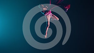 Young female circus gymnast spins hanging upside down on aerial silk. Acrobat performs tricks at height, red silk