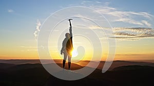 A young female climber victoriously raises her hands up while standing on the top of the mountain at sunset.