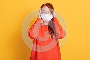 Young female with closed eyes touching her head, red haired woman wearing orange sweater and medical protective mask, standing