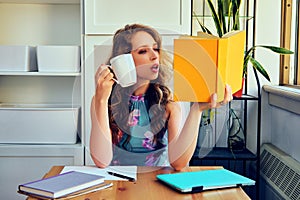 young female college student with book and cup of coffee studying at home reading homework