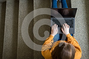 Young female college student sitting on stairs at school, writing essay on her laptop. Education concept.
