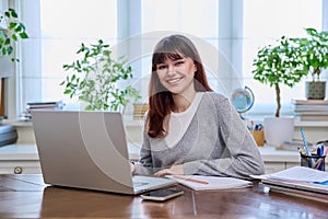 Young female college student studying at home using laptop, looking at camera