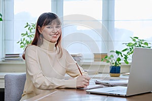 Young female college student studying at home using laptop, looking at camera