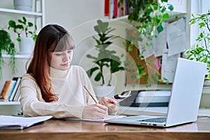 Young female college student studying at home using laptop, writing in notebook