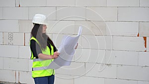 Young female construction worker in green high visibility vest and white hard hat, holding paper drawings with building