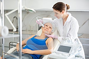 Young female cosmetologist doing microcurrent procedure to adult woman