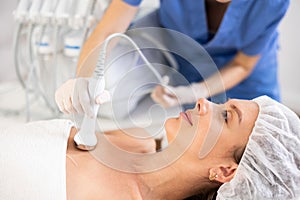 Young female cosmetologist performing ultrasound procedure on collarbones of young woman