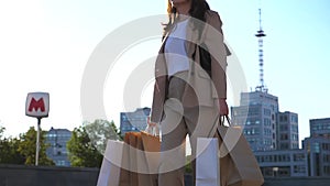 Young female customer holds shopping bags walking at sidewalk after purchases. Fashionable woman carries paper packets