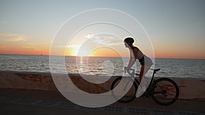 Young female is cycling on bicycle along sea promenade at sunrise. Female riding. Stylish attractive woman is pedaling on bike on