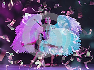 Young female dancers with angel`s wings in neon light on black background in flying confetti