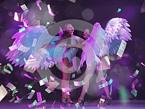 Young female dancers with angel`s wings in neon light on black background in flying confetti