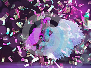 Young female dancers with angel`s wings in neon light on black background in flying confetti