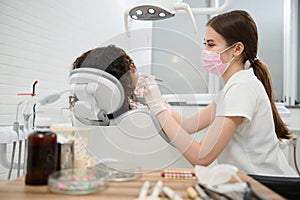 Young female dentist hygienist in protective medical mask and gloves performing oral treatment in contemporary dental clinic.