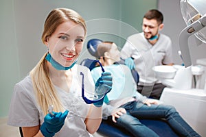 Young female dentist look on camera and smile. She hold glasses. Male dentist stand beside girl. Child sit in dental