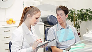 Young female dentist looking at digital tablet and listening male patient in dental clinic