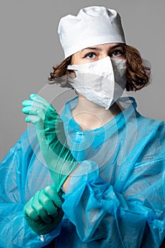 A young female doctor in a blue protective disposable coat and white face mask puts on latex gloves. Gray background