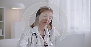 Young female doctor is communicating with patient online by laptop, using videocall and headphones with mic