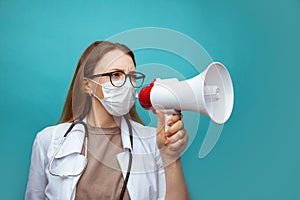 Young female doctor in face mask shouting into megaphone on blue background