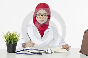 Doctor with hijab, sitting and writing something in her book over white background