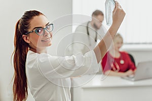 Young female doctor in hospital looking at x-ray. The other two doctors communicate in the background.